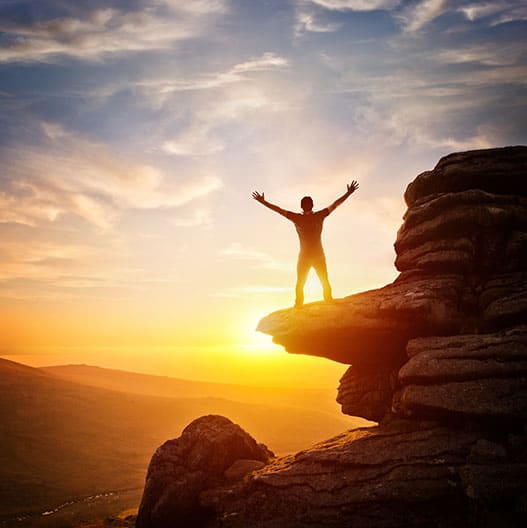 Guy standing on a cliff in front of sunset stretching his hands to the sky