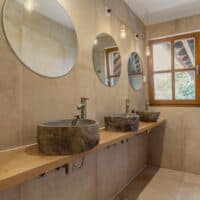 A modern bathroom features stone sinks, round mirrors, hanging light bulbs, and a wooden window overlooking a green outdoor area. Neutral tone tiles adorn walls.
