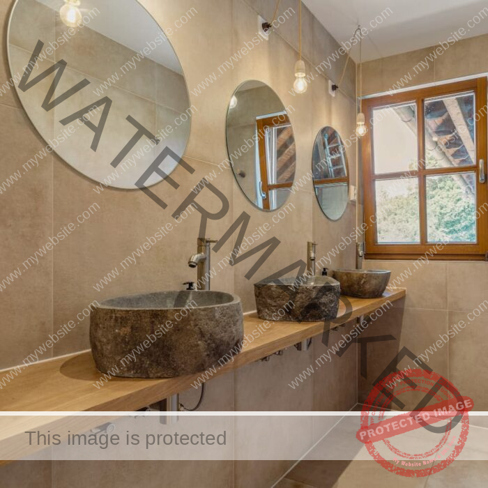 A modern bathroom features stone sinks, round mirrors, hanging light bulbs, and a wooden window overlooking a green outdoor area. Neutral tone tiles adorn walls.