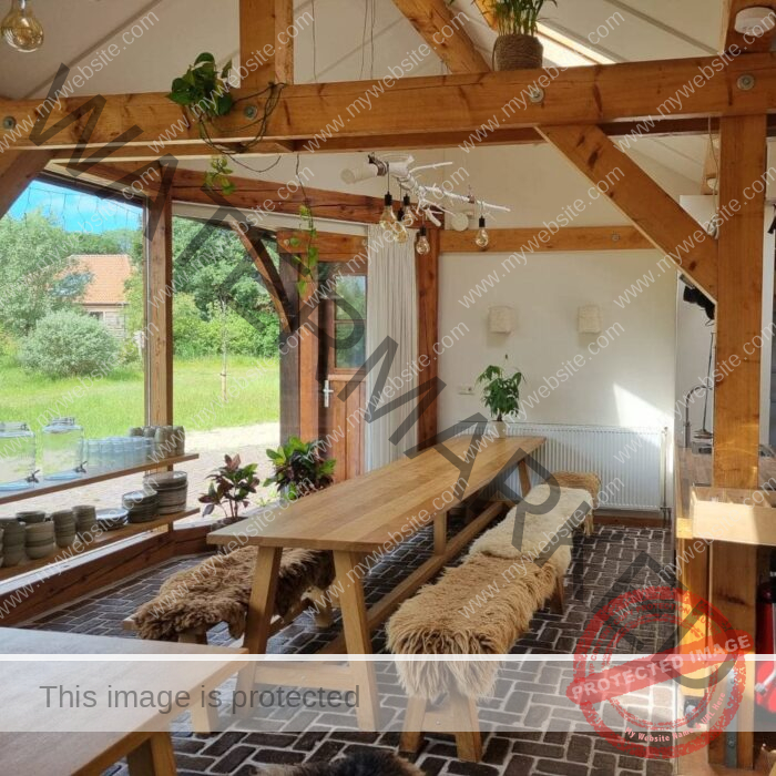 A cozy, rustic wooden dining area with large windows overlooking a green yard, featuring long tables, sheepskin-covered benches, and potted plants.