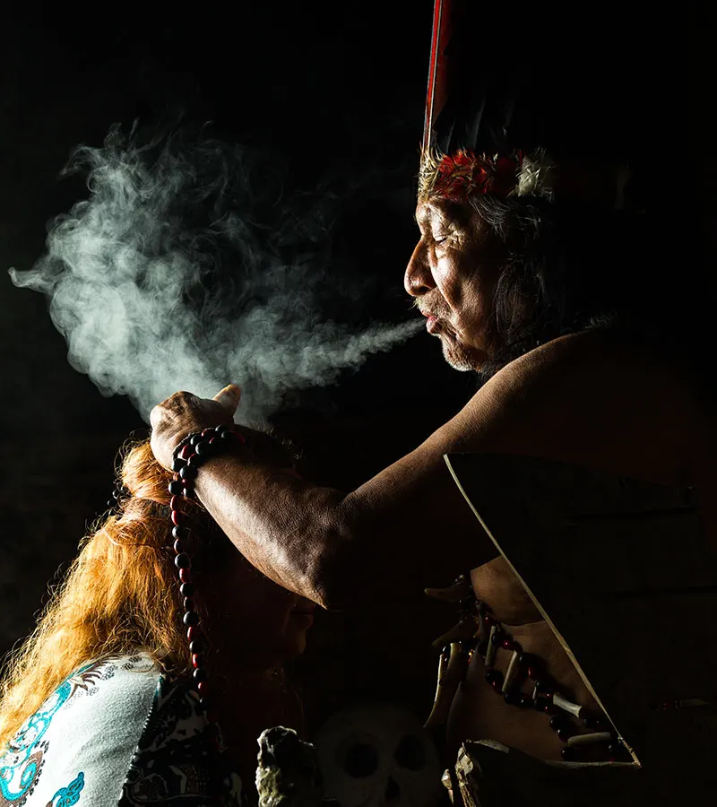 Shaman blows smoke over head of a woman, background is black