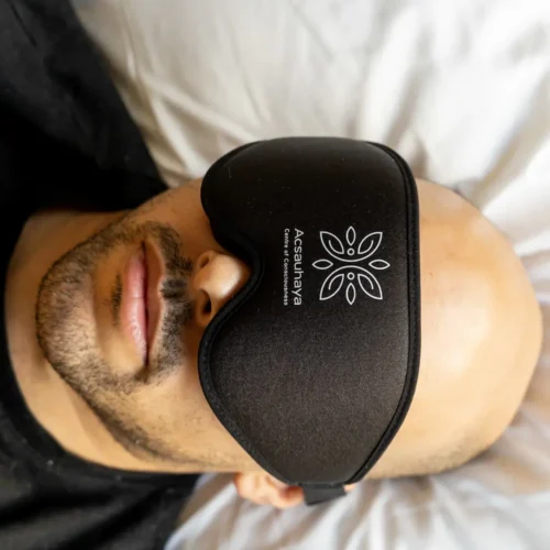 A person with a bald head and beard is lying on a bed, wearing a black sleep mask with the logo and text "Acsauhaya" printed on it. The background shows a white pillow. Picture taken during a psilocybin session.