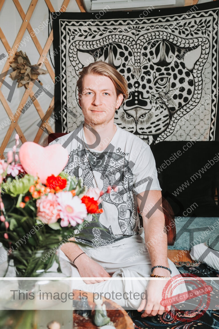 Young blonde guy, sitting in white clothes on the ground, flowers is the foreground and a jaguar painting in the background