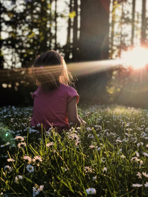 Little girl sitting on grass, sun shines through forest