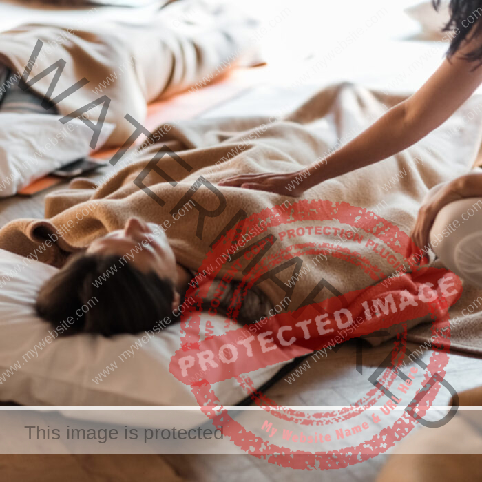 Woman laying on mattress with brown blanket, another woman touching her chest