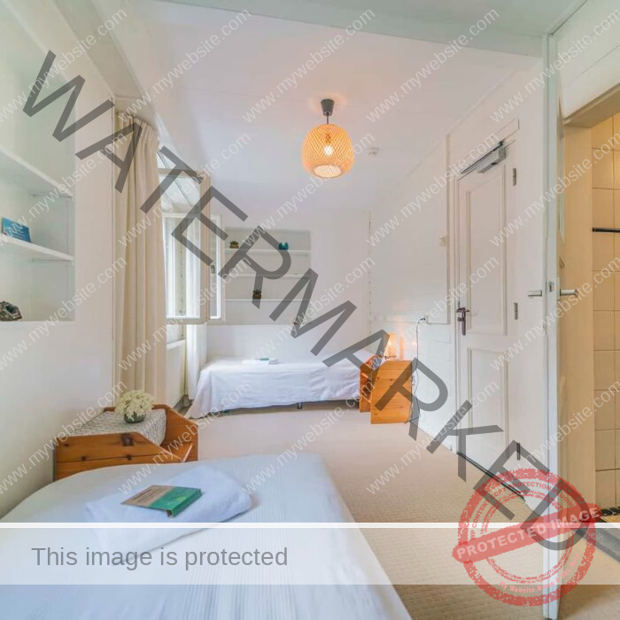 A bright, minimalist bedroom with two single beds adorned with white linens. A wooden nightstand with a lit lamp sits between them. Shelves are on the opposite wall, and a window lets in natural light. The room has a door leading to a small tiled bathroom.