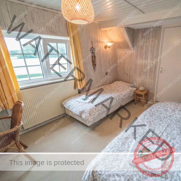 A cozy bedroom with two single beds covered in grey and white patterned bedspreads. A window with yellow curtains lets in natural light. The room has wooden walls, a rattan chair, and a small bedside table, creating a warm and inviting atmosphere.