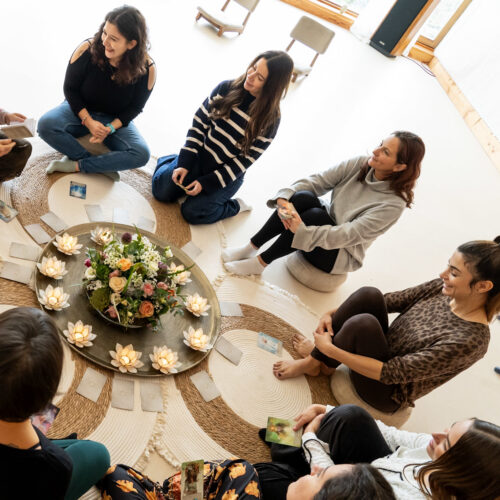 Women sit in a circle on the floor around flower arrangements and candles, each holding cards. The atmosphere appears calm and focused, at a psilocybin retreat.