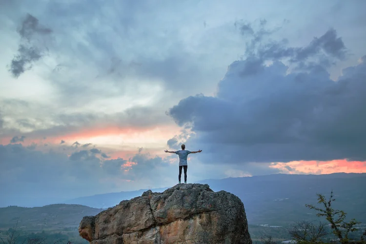 Man standing on rock, arms wide open to the sky, cloudy sunset