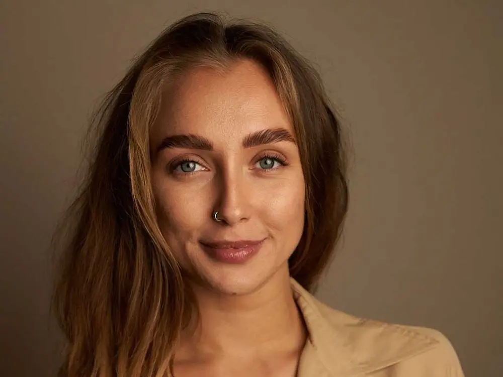 Portrait of Anastasiya, a young woman with long brown hair, arched eyebrows, and a nose ring smiles softly at the camera. She is wearing a beige top and is posed against a plain, neutral background.