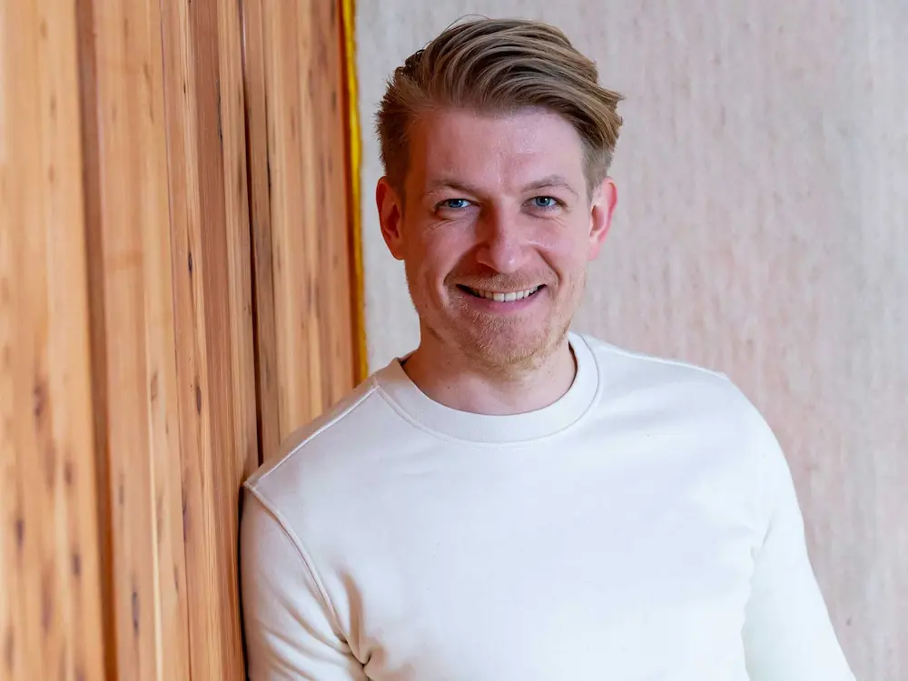 Portrait of Joe, a smiling man with light hair wearing a white long-sleeve shirt stands against a wooden wall, looking directly at the camera.