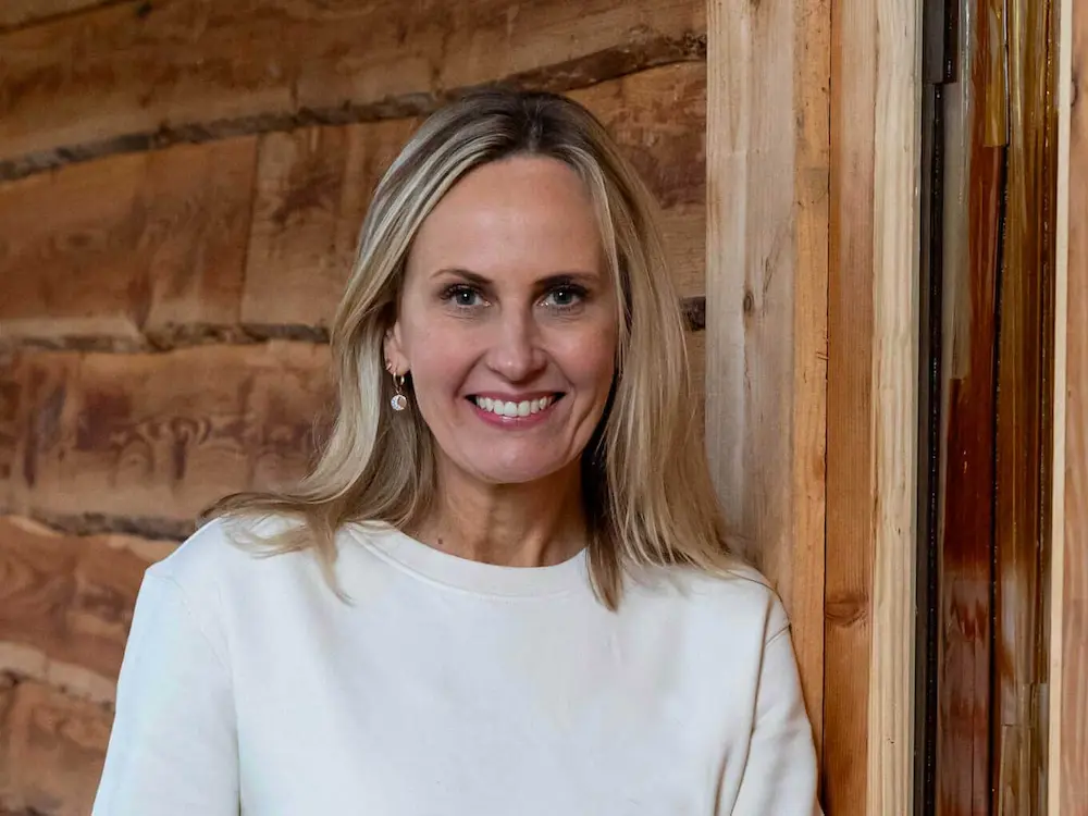 Portrait of Magda: A woman with long blonde hair, wearing a white top and drop earrings, stands smiling in front of a wooden wall with visible logs and a door frame.