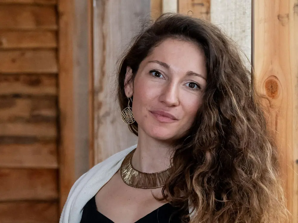 Portrait of Maria, a woman with long, curly brown hair wearing a white shawl, gold statement necklace, and gold earring, stands indoors against a wooden wall, looking at the camera with a slight smile.