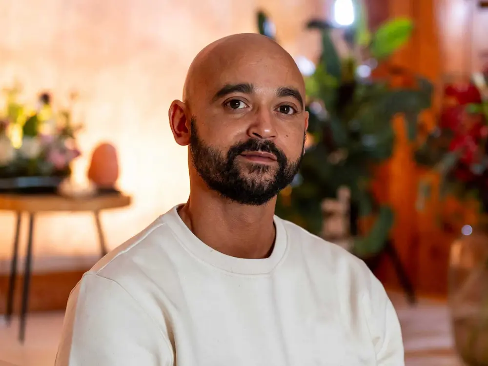 Portrait of Shurandy, a man with a shaved head and beard wears a cream-colored sweatshirt and sits indoors, surrounded by blurred plants and warm lighting in the background.