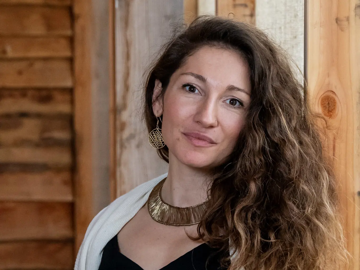 A woman with long curly brown hair, wearing a gold necklace, gold earrings, and a white shawl over a black top, stands in front of a wooden wall, looking calmly at the camera.
