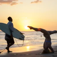 Sunset at the beach, a woman doing yoga and a guy with his surfboard in the foreground