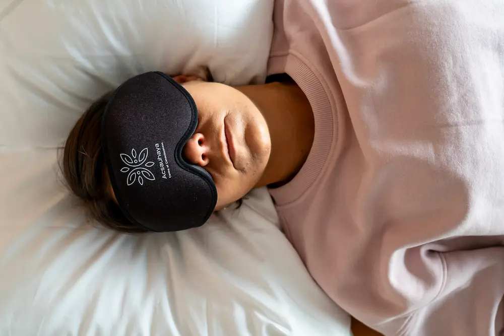 Eine Person, die eine schwarze Schlafmaske und ein hellrosa Sweatshirt trägt, liegt auf weißen Laken und scheint sich während einer Psilocybin-Zeremonie auszuruhen oder zu schlafen.