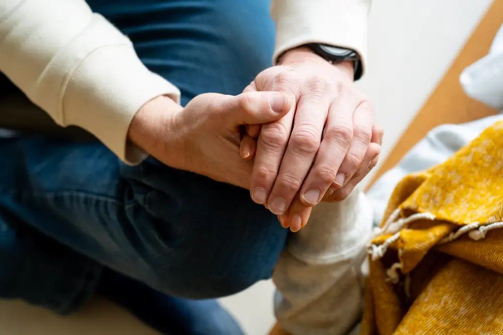 Nahaufnahme einer Person, die sanft die Hand einer anderen Person hält und ihr Trost und Unterstützung bietet. Die Szene vermittelt Empathie und Fürsorge und erinnert an die Wärme, die oft in Psilocybin-Therapiesitzungen gesucht wird. Beide Personen tragen Freizeitkleidung.