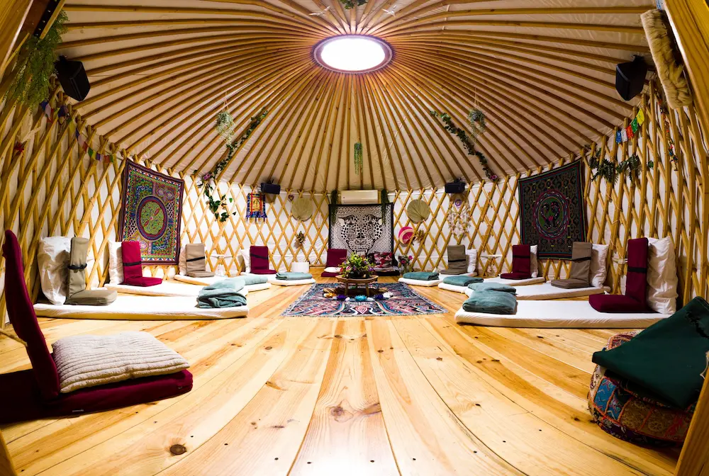 Das Innere einer Jurte verfügt über einen Holzboden, ein rundes Oberlicht, Bodenkissen, gefaltete Decken und bunte Wandteppiche, die an den Wänden hängen und eine gemütliche und einladende Atmosphäre schaffen.