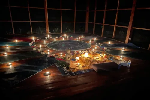 A dimly lit ayahuasca ceremony space with candles arranged in a circle around a central altar decorated with a small statue, flowers, and cloths, creating a serene, ritualistic atmosphere.