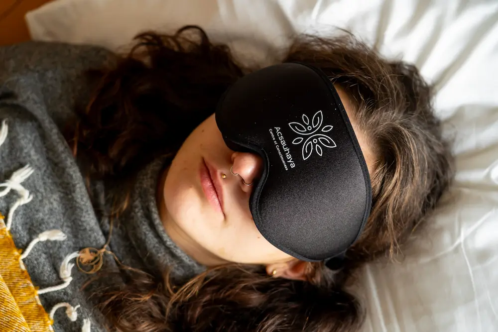 A person with long brown hair lies on a bed wearing a gray sweater and a large black sleep mask with the Acsauhaya logo printed on it. The person appears to be resting or sleeping.