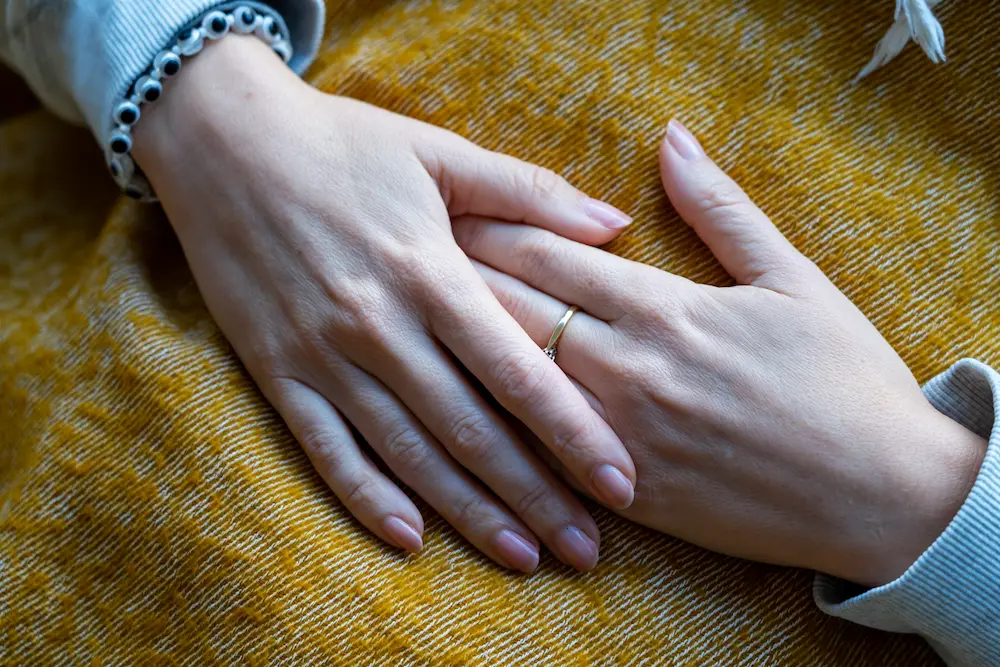 Close-up of two hands resting on a yellow textured fabric; one hand wears a simple gold ring on the ring finger and a beaded bracelet on the wrist.