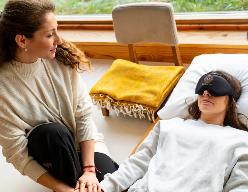 Eine Frau sitzt neben einer anderen Frau, die mit einer Augenmaske liegt, und hält ihr während eines Psilocybin-Retreats unterstützend die Hand. Die Szene wirkt ruhig und beruhigend, im Hintergrund sind eine Decke und ein Stuhl zu sehen.