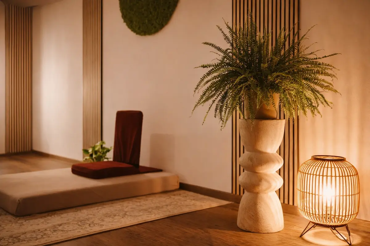 A serene meditation room with a cushion and backrest on a mat, a tall potted fern on a sculpted stand, and a glowing rattan lamp, all set against warm-toned walls and wooden floors.