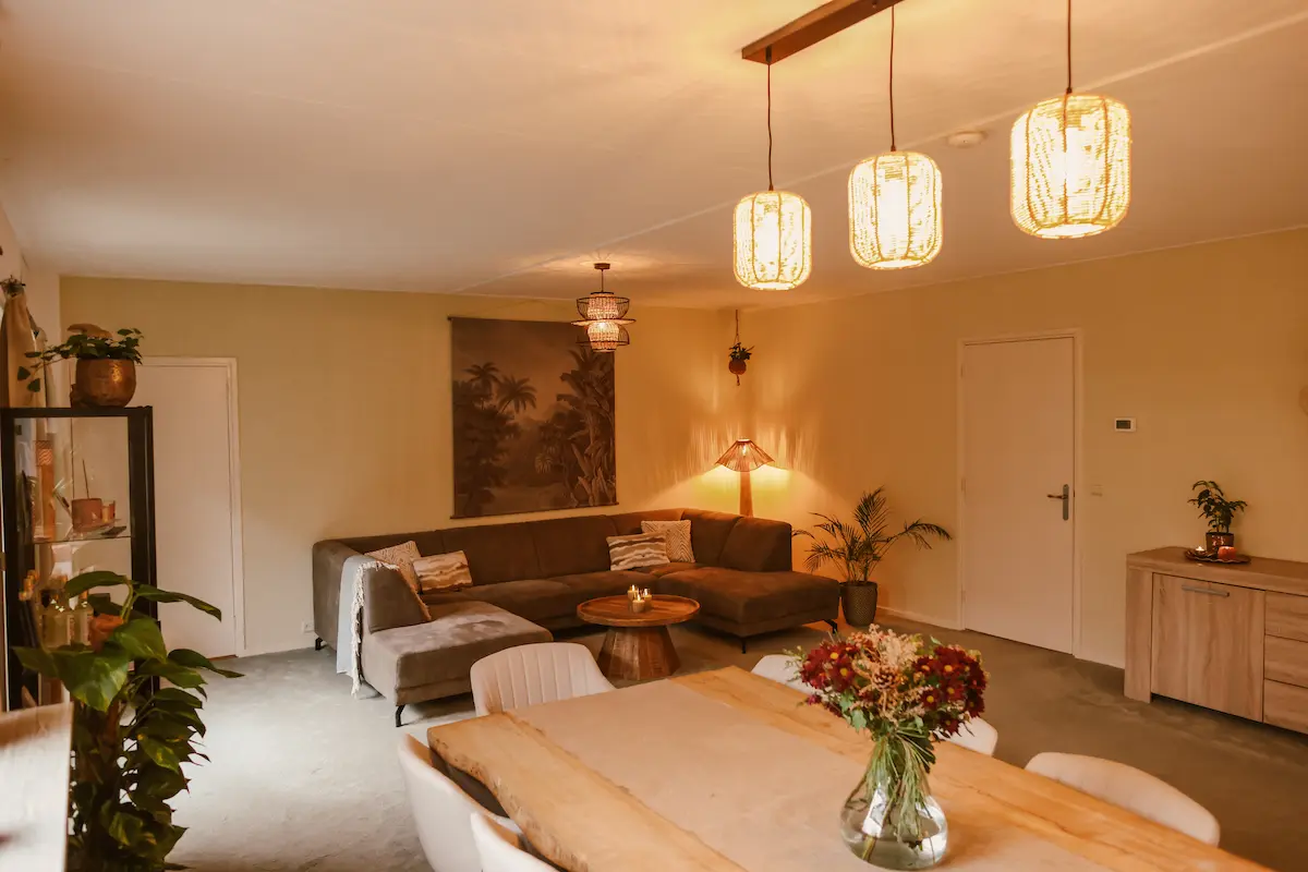 A warm, inviting lounge with a large corner sofa, wooden coffee table, dining table with flowers, indoor plants, and pendant lights creating a cosy atmosphere. Neutral tones and soft lighting dominate the décor.