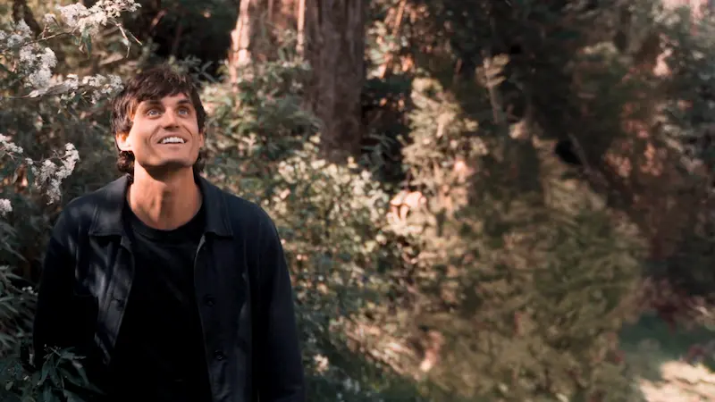 A man with dark hair wearing a black shirt and jacket stands outdoors among greenery, smiling and looking upward. Sunlight filters through the trees in the background.