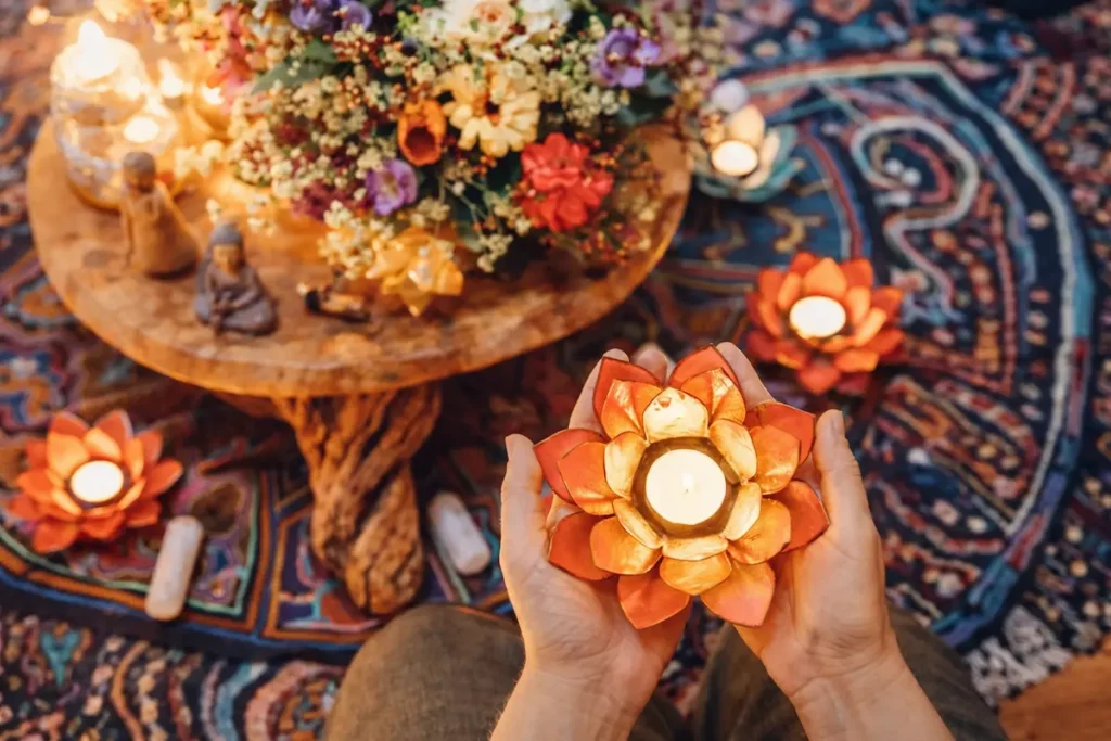 Hands hold a lotus-shaped candle holder with a lit tealight on a colourful patterned rug, evoking an ayahuasca ceremony. Nearby, other candles, crystals, and a round wooden table with flowers create a warm and peaceful scene.