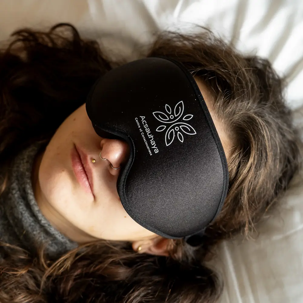 1 or 2 Day Private Ayahuasca Retreat A person with long curly hair wears a black eye mask labeled "Acsauhaya" while lying on a white pillow, during a ayahuasca retreat session.
