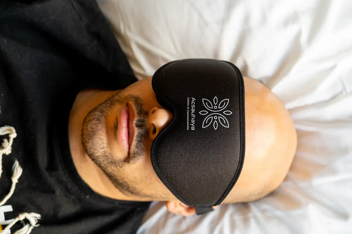 A man with a beard and mustache lies on a white pillow, wearing a large black sleep mask with a white Acsauhaya logo during a psilocybin retreat session.