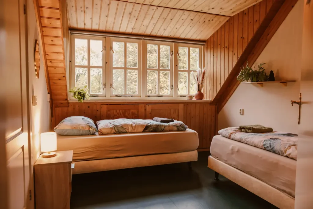 Cozy bedroom with wooden walls and ceiling, two single beds with floral bedding, a nightstand with a lamp, and large windows letting in natural light. Plants and decorative items are on a shelf and windowsill.