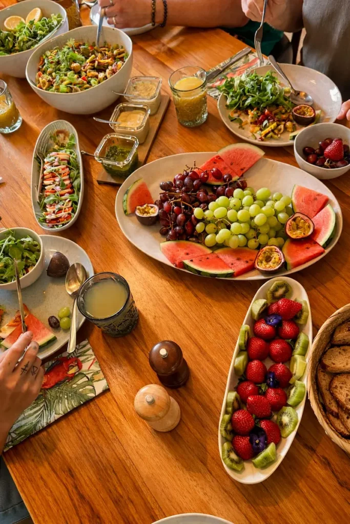 A wooden table set with bowls and plates of fresh fruit, salad, bread, and drinks. People with forks are reaching for food, enjoying a colourful, healthy meal together.