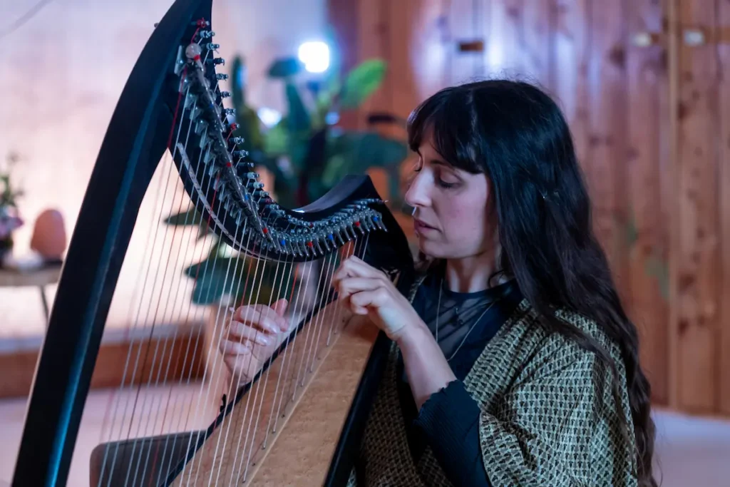Fralalai A woman with long dark hair plays a harp indoors, focusing intently on the strings. The background features soft lighting, plants, and wooden walls, creating a warm and cozy atmosphere.