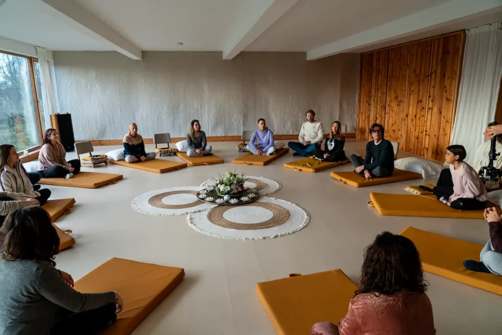 Psilocybin session A group of people sit in a circle on yellow mats in a bright room, surrounding a decorative centerpiece with flowers, participating in a psilocybin retreat.
