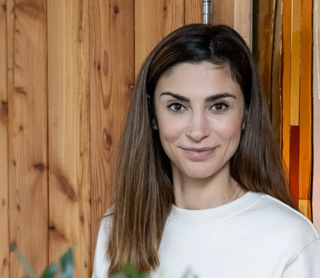 Sofia A woman with straight brown hair and a white sweater stands indoors in front of wooden paneling and stained glass, smiling softly at the camera. Some green leaves are visible in the foreground.