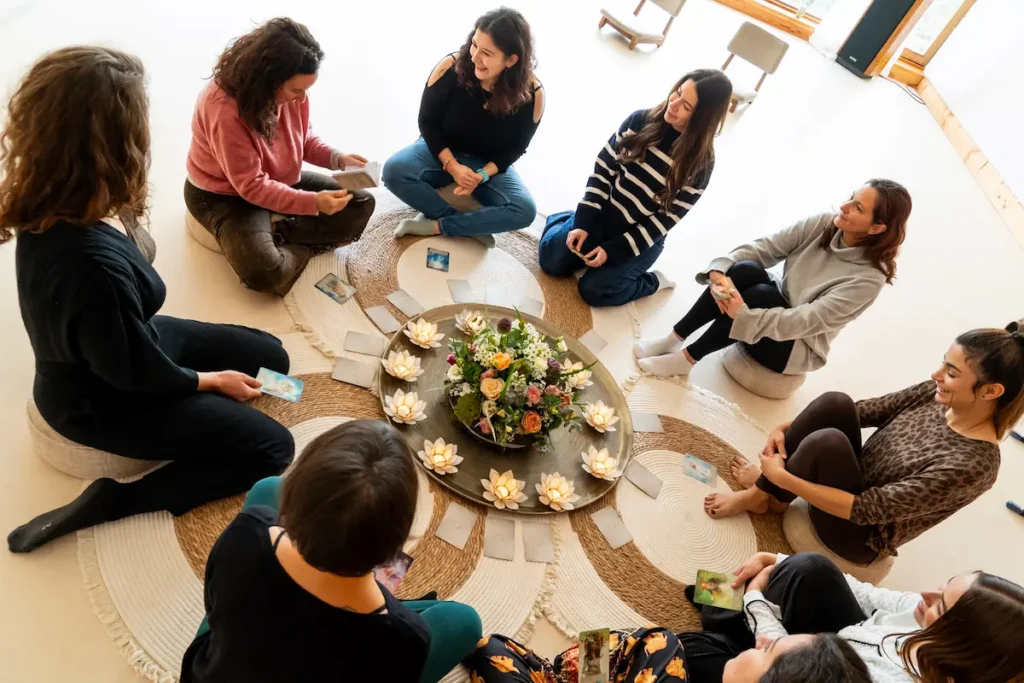 Women psilocybin retreat Eight women sit in a circle on the floor around flower arrangements and candles, each holding cards. The atmosphere appears calm and focused, at a psilocybin retreat.