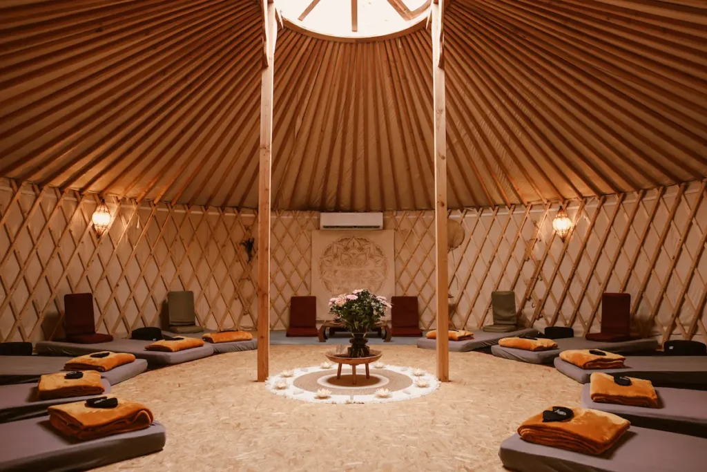 Interior of a round yurt with wooden beams and soft lighting, featuring cushions, folded blankets, and eye masks arranged in a circle around a central table adorned with flowers and candles.